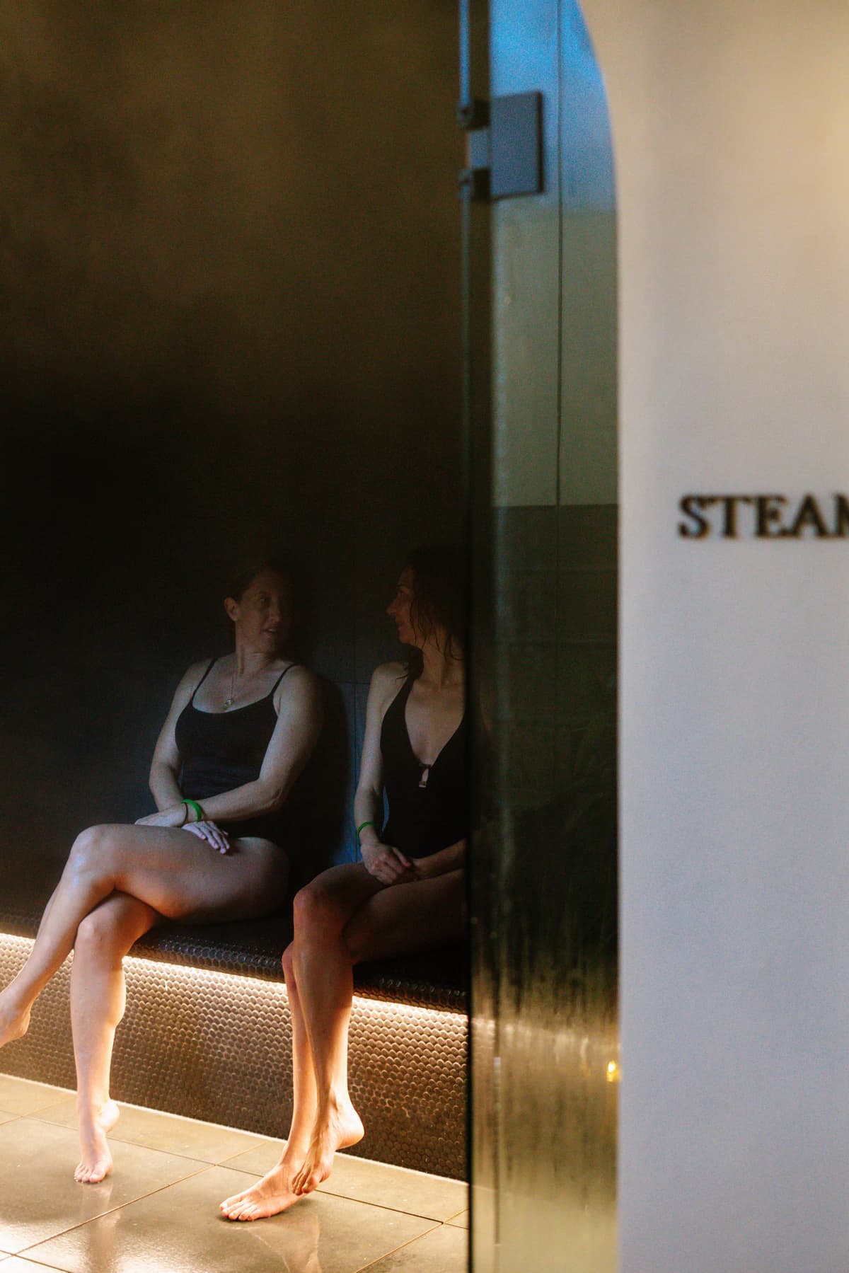 Two friends talking on the warmly lit bench of the Navia steam room, seen through the glass door