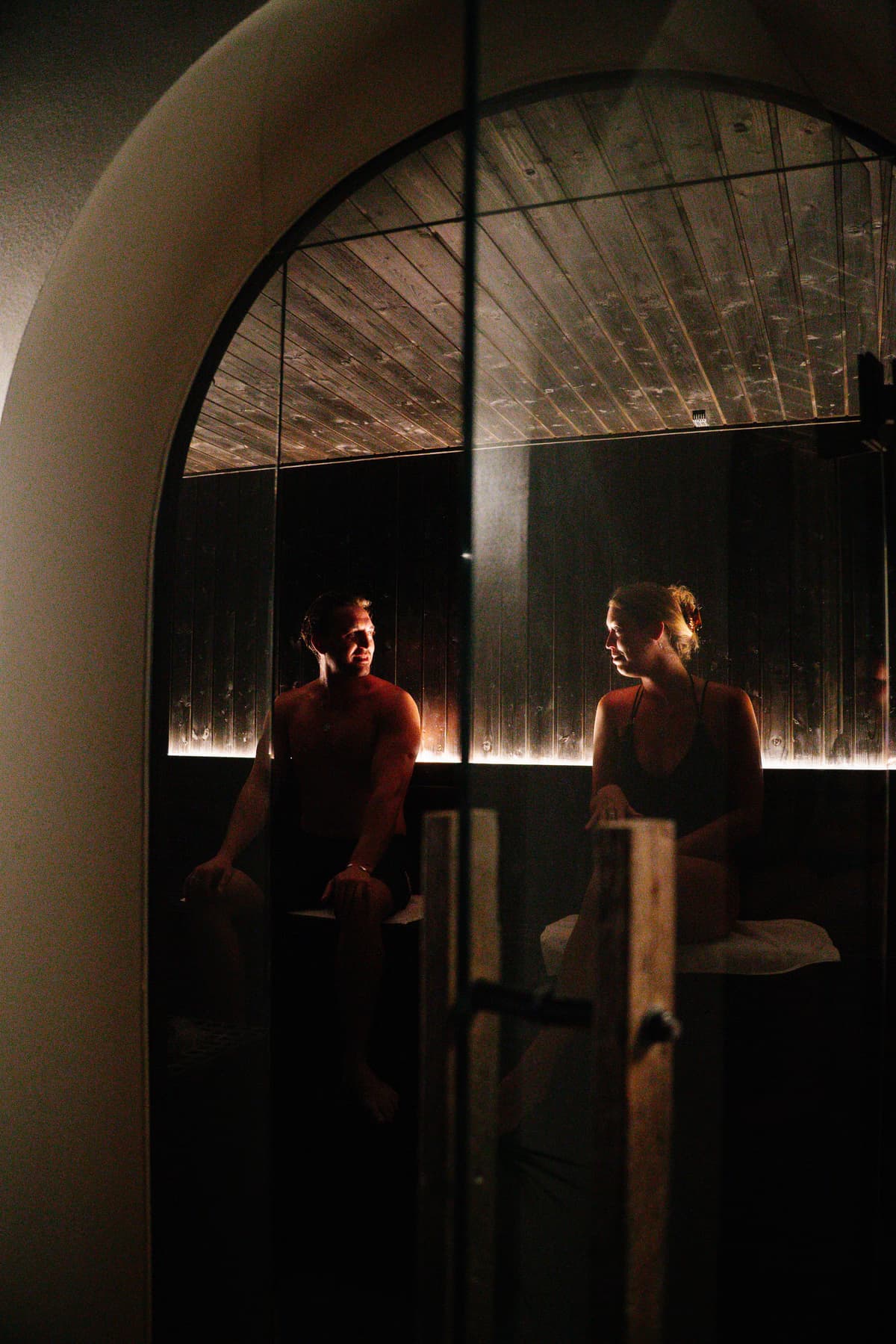 Couple sharing the warm sauna, seen through the arched glass door at Navia Bathhouse