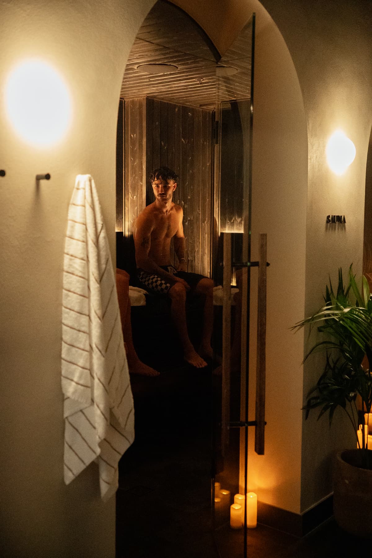 A bather seated in the warm-lit timber sauna at Navia Bathhouse, viewed through an arched doorway lit by candles