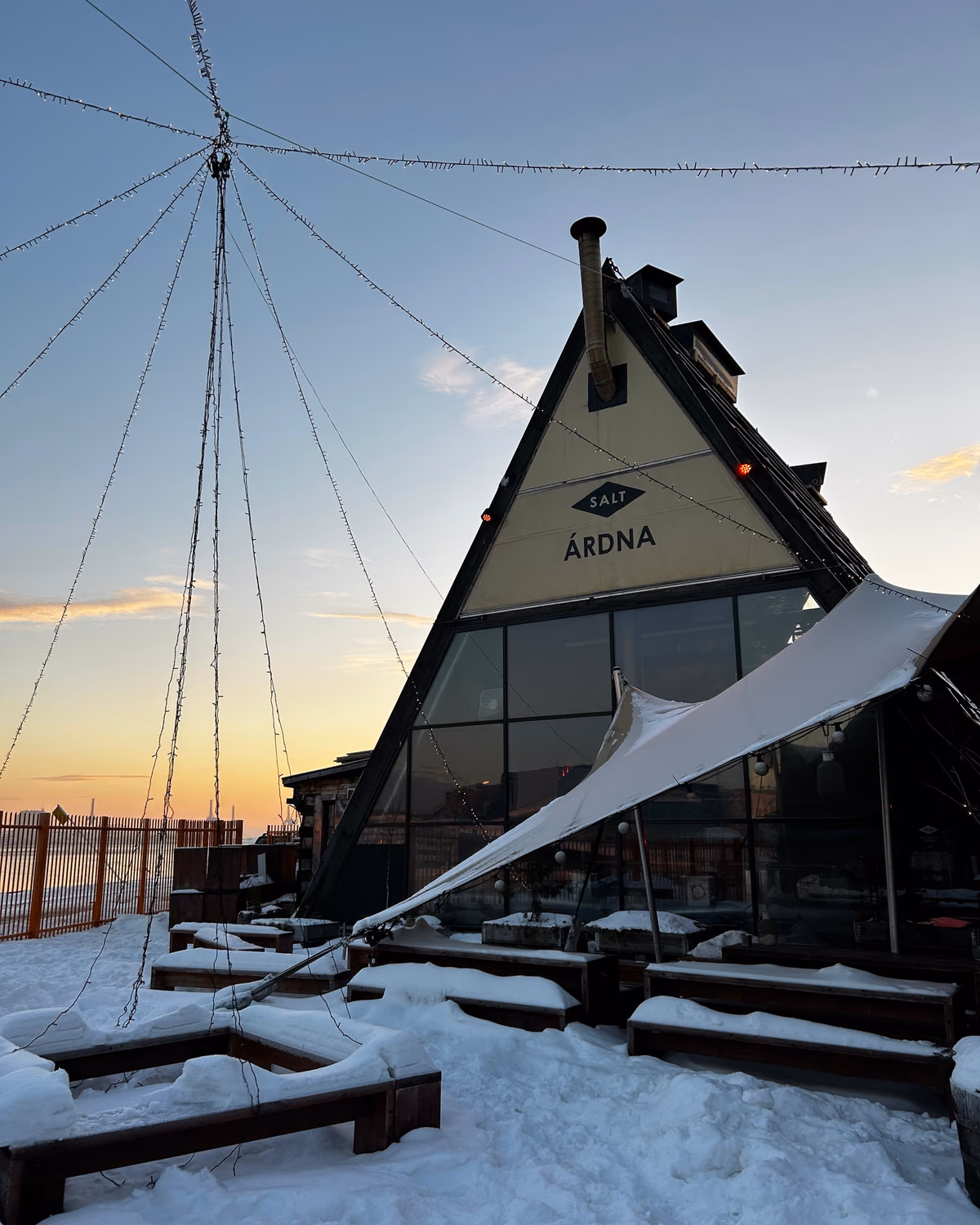 The A-frame timber sauna of SALT Ardna in Oslo at dusk, snow on the ground and festoon lights strung overhead