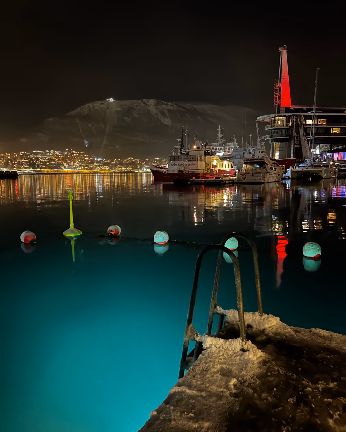 The sea pool at Pust in Tromso at night, turquoise water beside a snow-covered ladder with the lit harbour and mountain behind