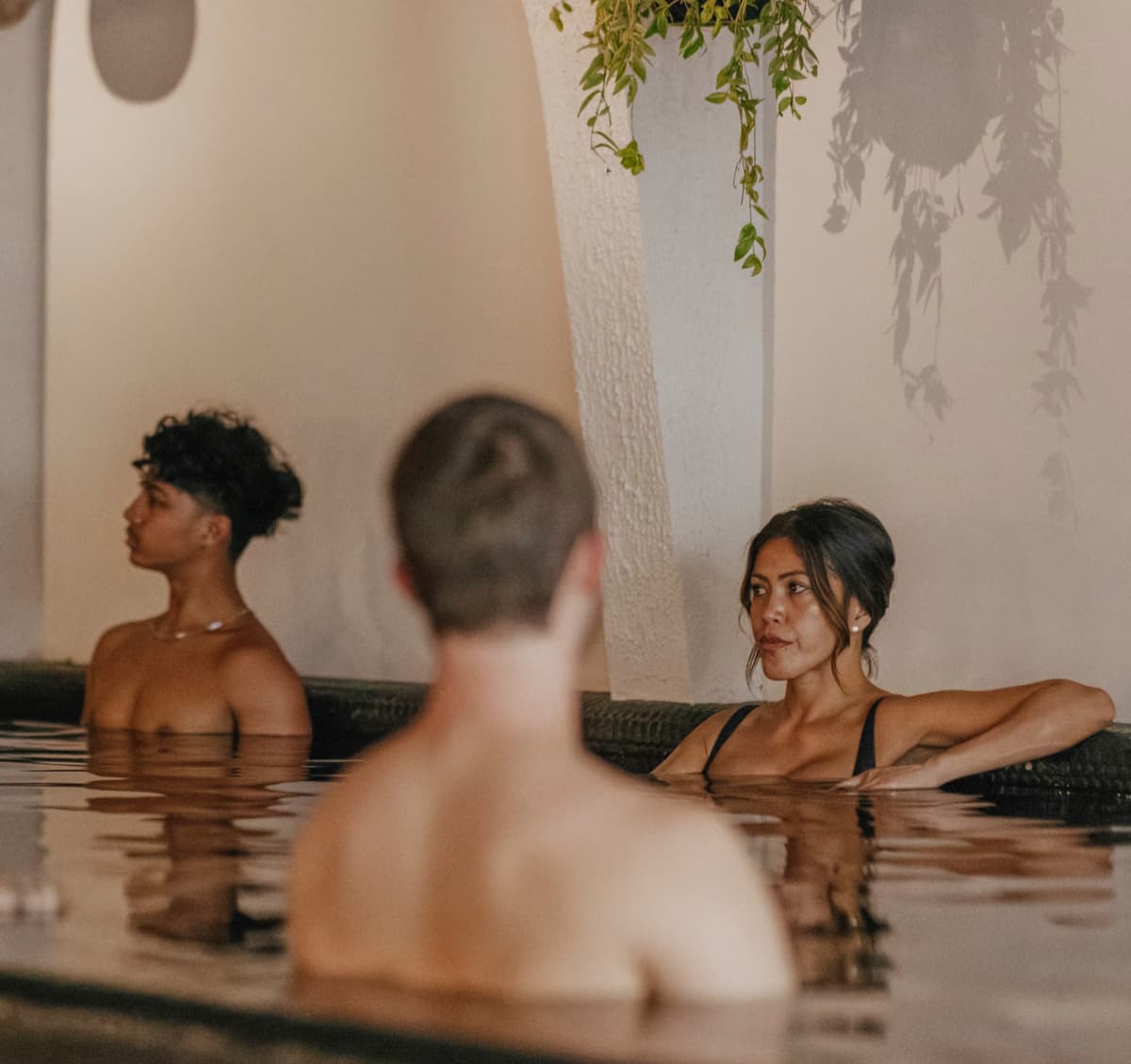 Three bathers together in the dim mineral pool at Navia Bathhouse beneath trailing plants