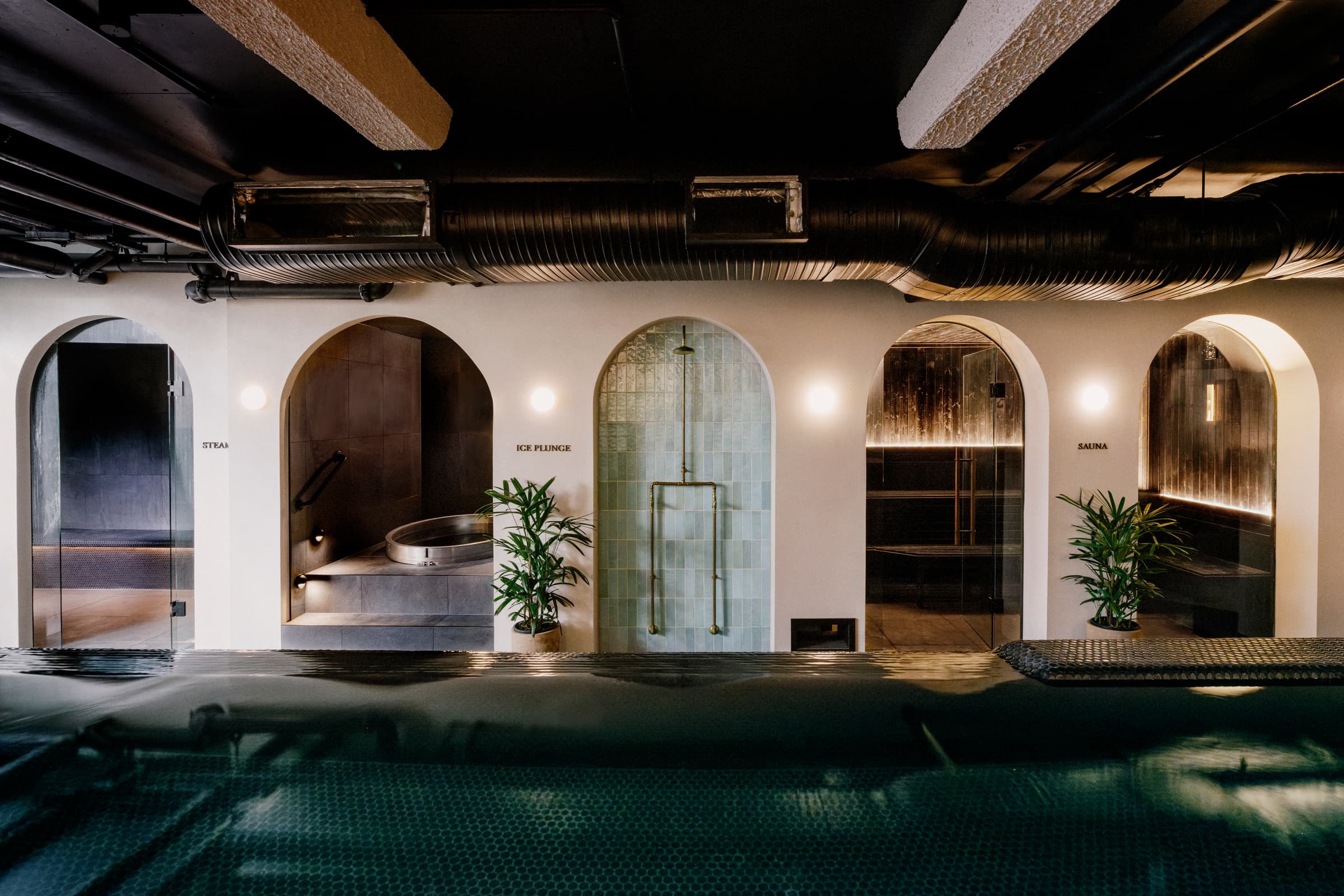 White-plaster arched alcoves housing steam room, ice plunge and sauna beside a dark-tiled thermal pool at Navia Bathhouse Byron Bay